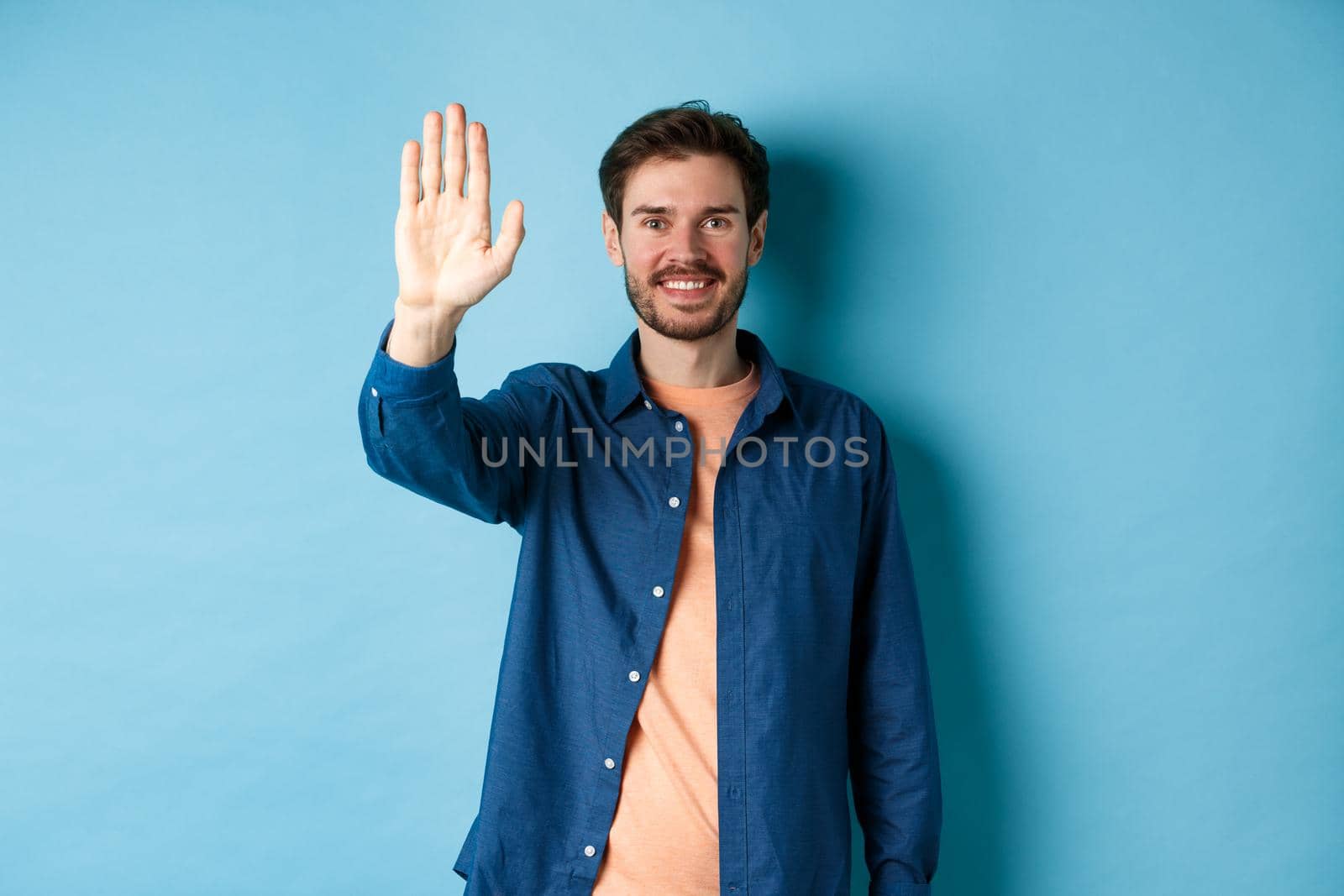 Friendly young man smiling and raising hand up for high five, waiving to greet or say hello, standing on blue background.