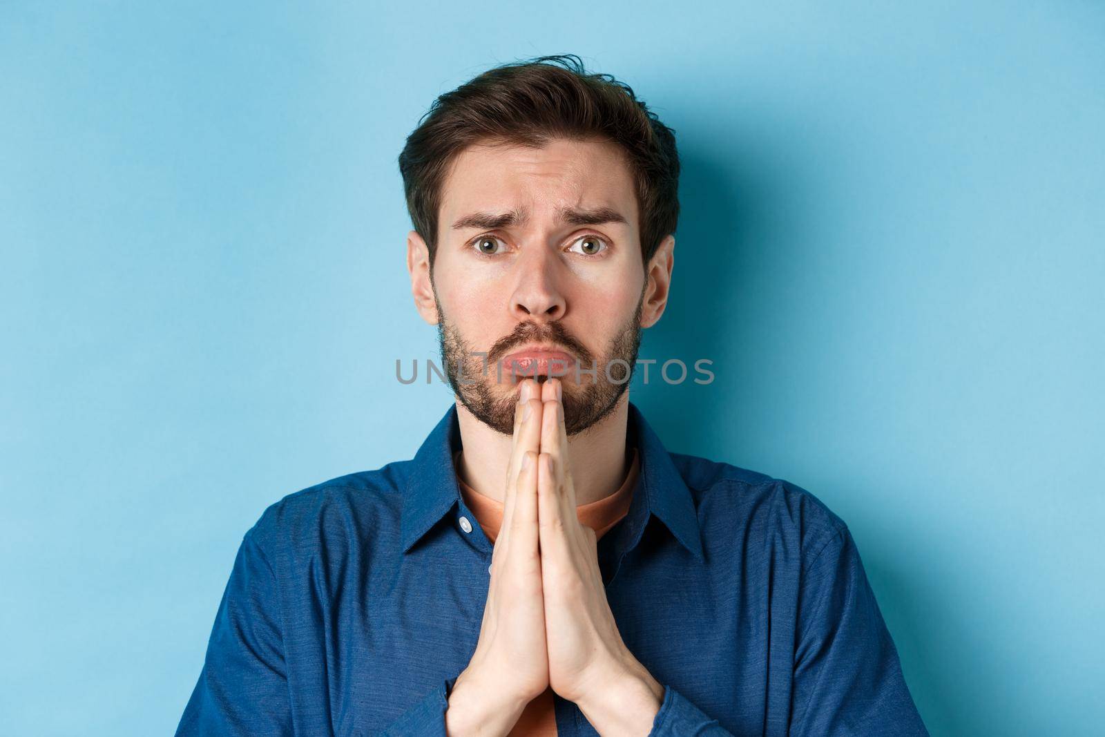 Sad young man begging for help, apoligizing and sobbing miserable, standing on blue background.