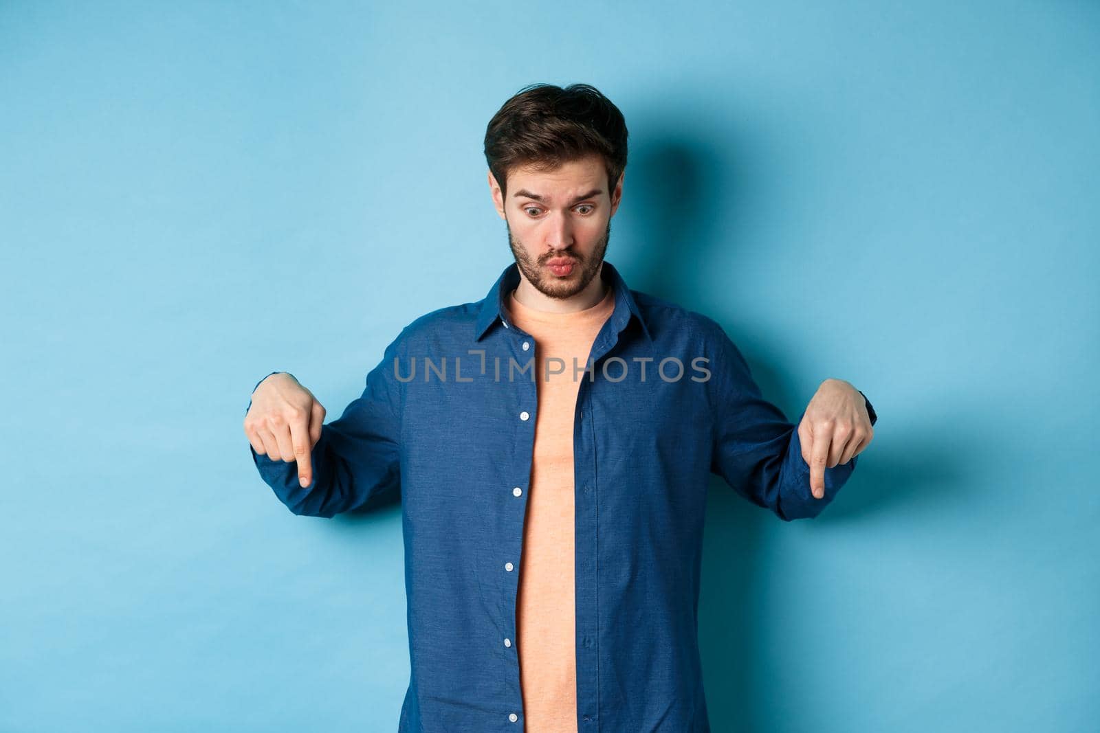 Image of shocked young man in casual clothes, looking and pointing down at something strange, standing on blue background.