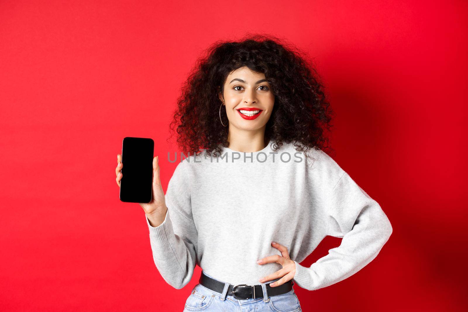 Attractive smiling woman showing empty smartphone screen and looking happy, advertising online store or application, standing against red background.