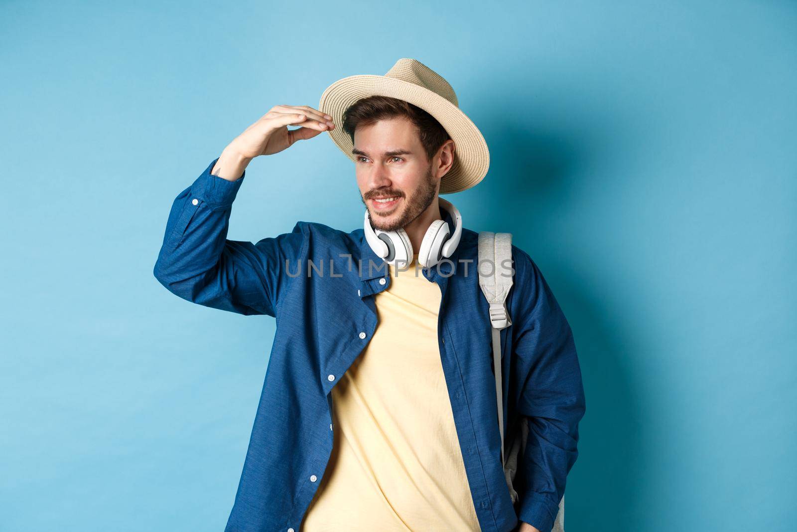 Handsome tourist in straw hat, covering face from sun and looking at distance aside, smiling pleased, travelling on summer vacation, standing over blue background.