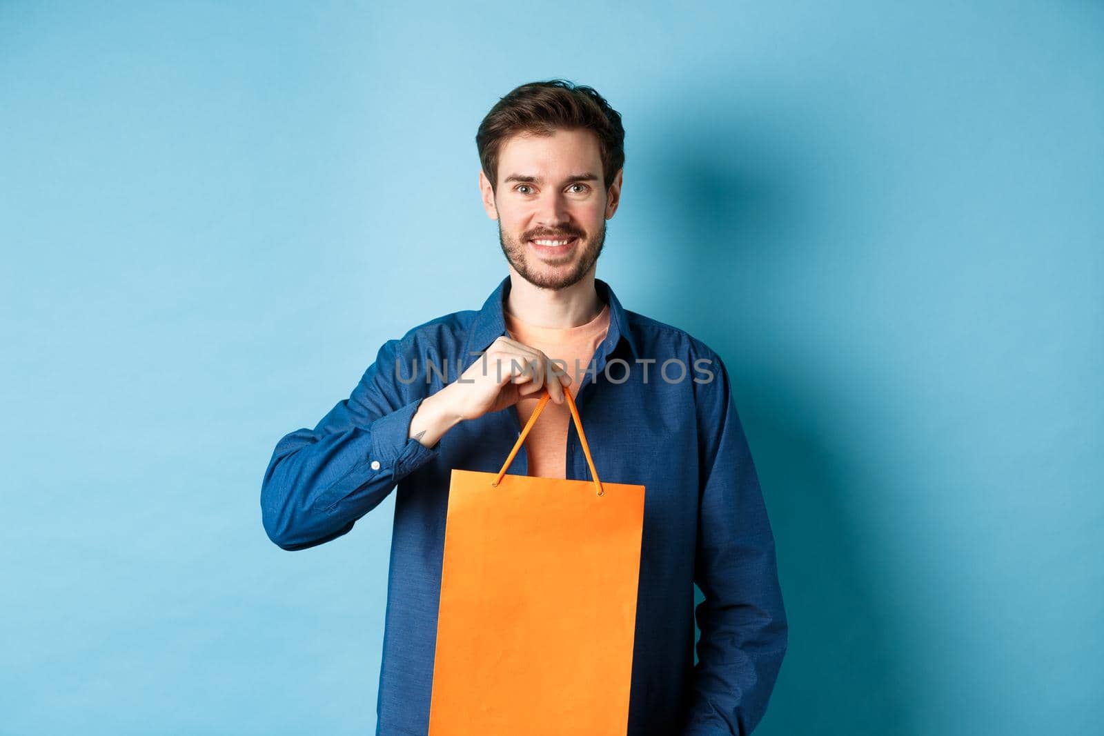 Handsome young man with beard, smiling and showing shopping bag, buying something in store, standing on blue background.