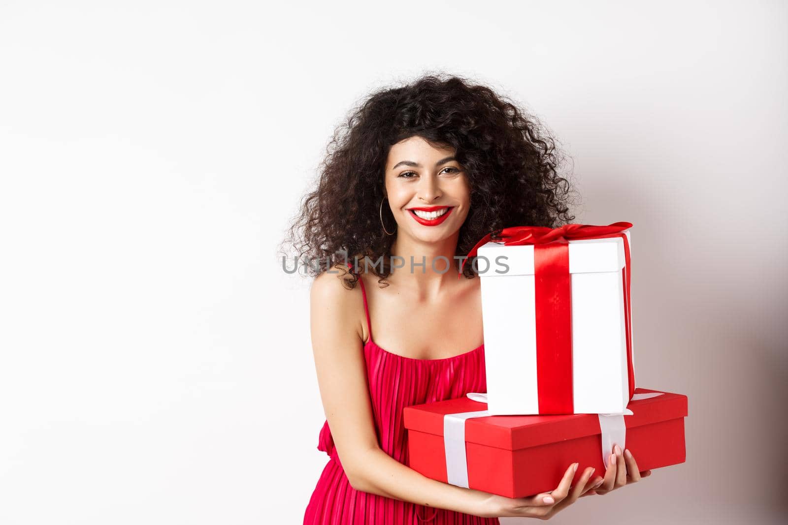Beautiful birthday girl with curly hair, holding bday gifts and smiling happy, celebrating, standing against white background.