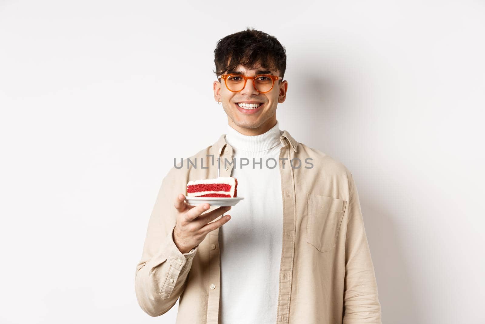 Happy caucasian guy celebrating birthday, holding cake with candle and smiling cheerful at camera, standing on white background.