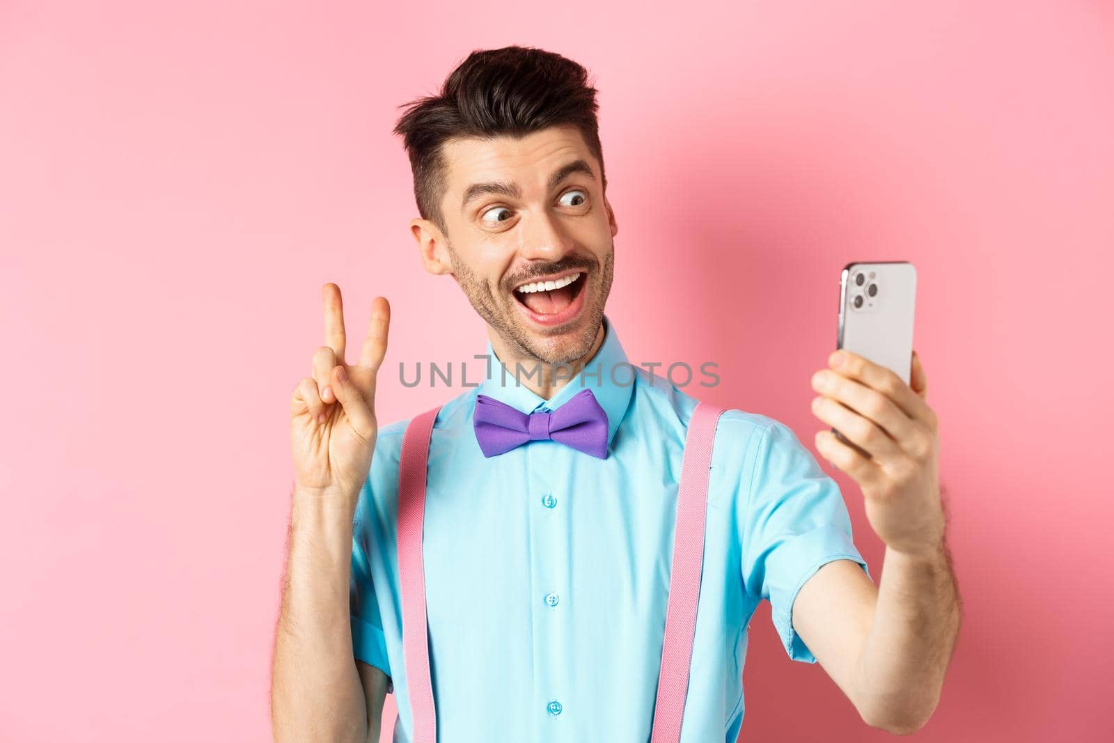 Technology concept. Funny man with moustache and bow-tie taking selfie on smartphone, showing peace sign and smiling at mobile camera, pink background.