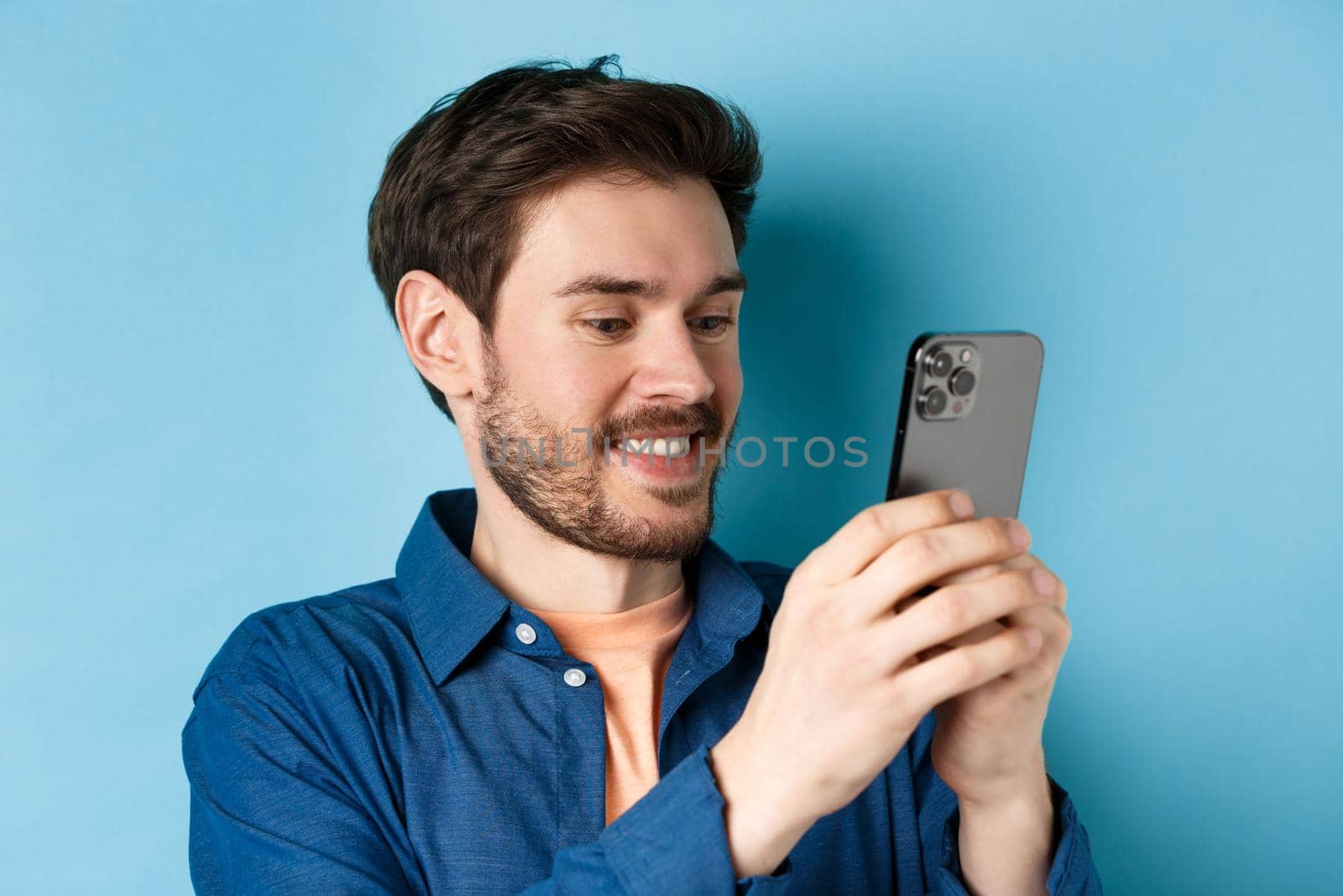 Close-up of handsome caucasian man writing message, reading mobile screen and smiling, standing on blue background by Benzoix