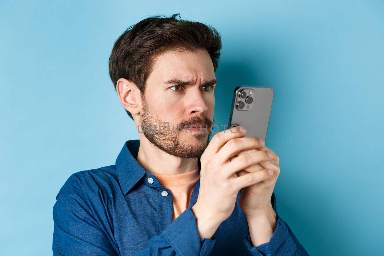 Confused guy staring close at mobile phone screen and frowning, standing on blue background.