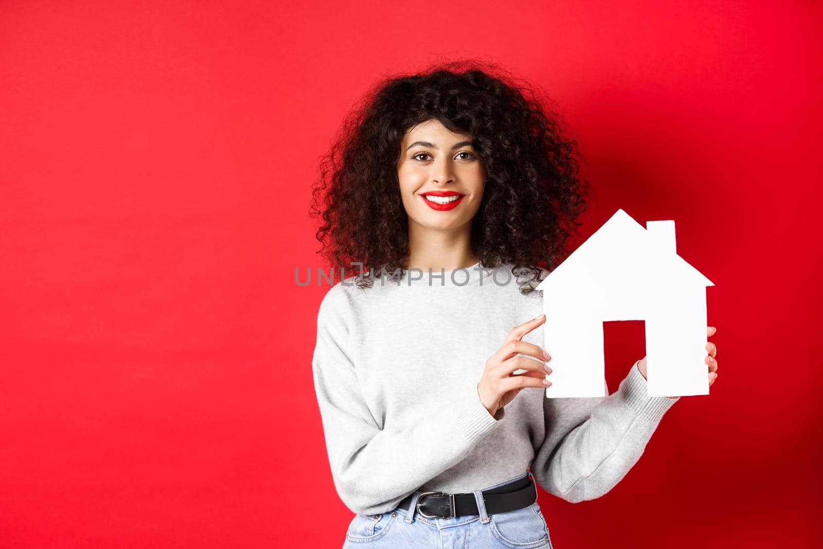 Real estate. Smiling caucasian woman with curly hair and red lips, showing paper house model, searching property, standing on red background.