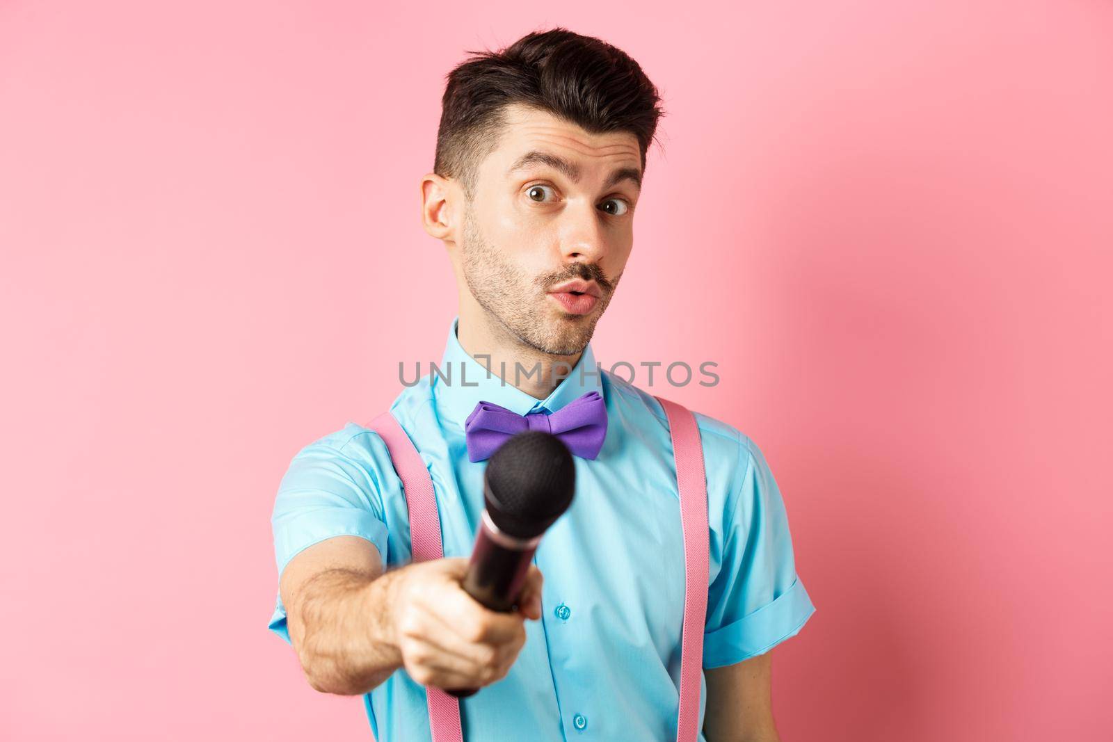 Handsome male show host stretch out hand with microphone, asking for interview or comments, standing on pink background.