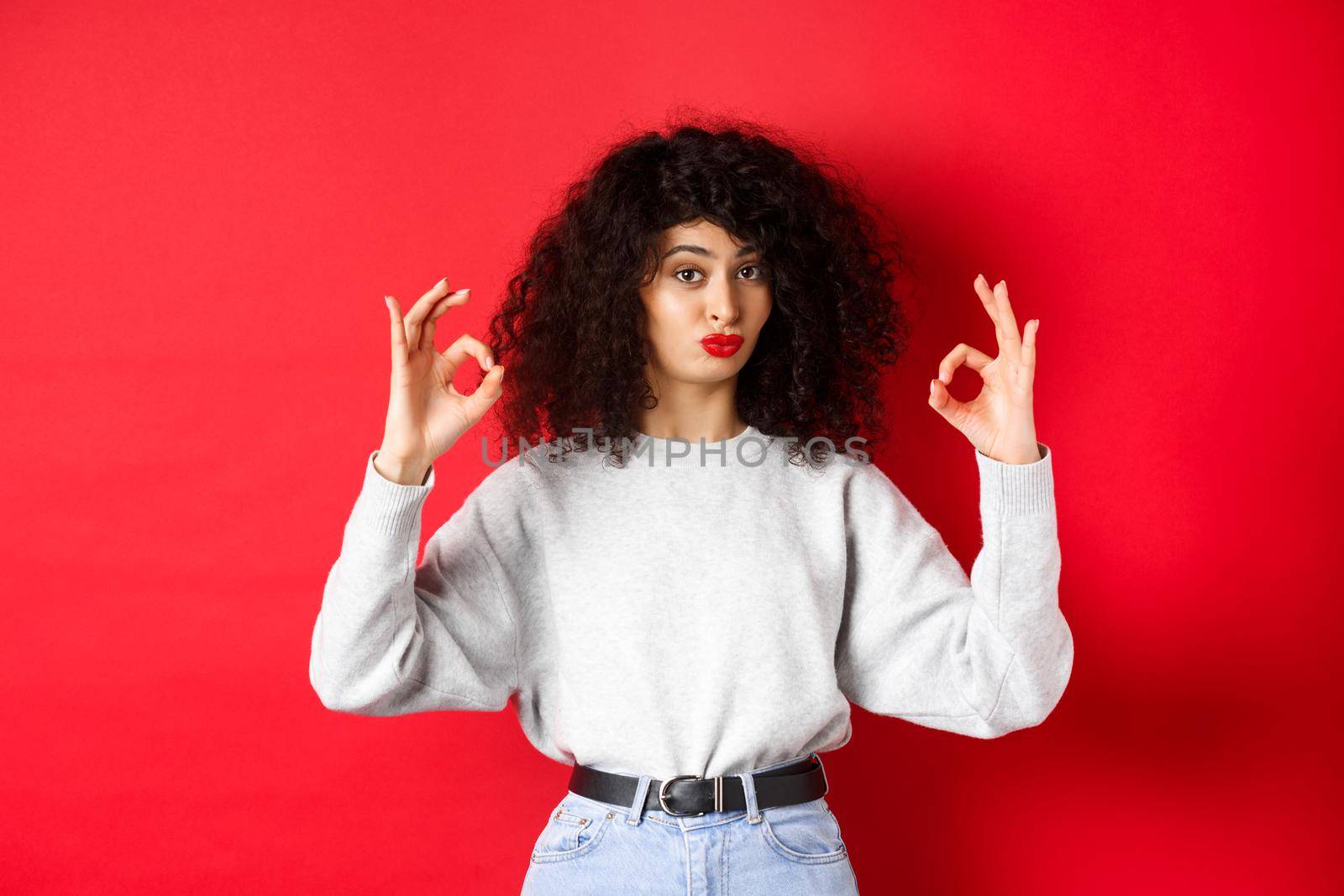 Not bad. Impressed curly woman nod in approval, pucker lips and showing okay signs, like good thing, standing satisfied on red background.