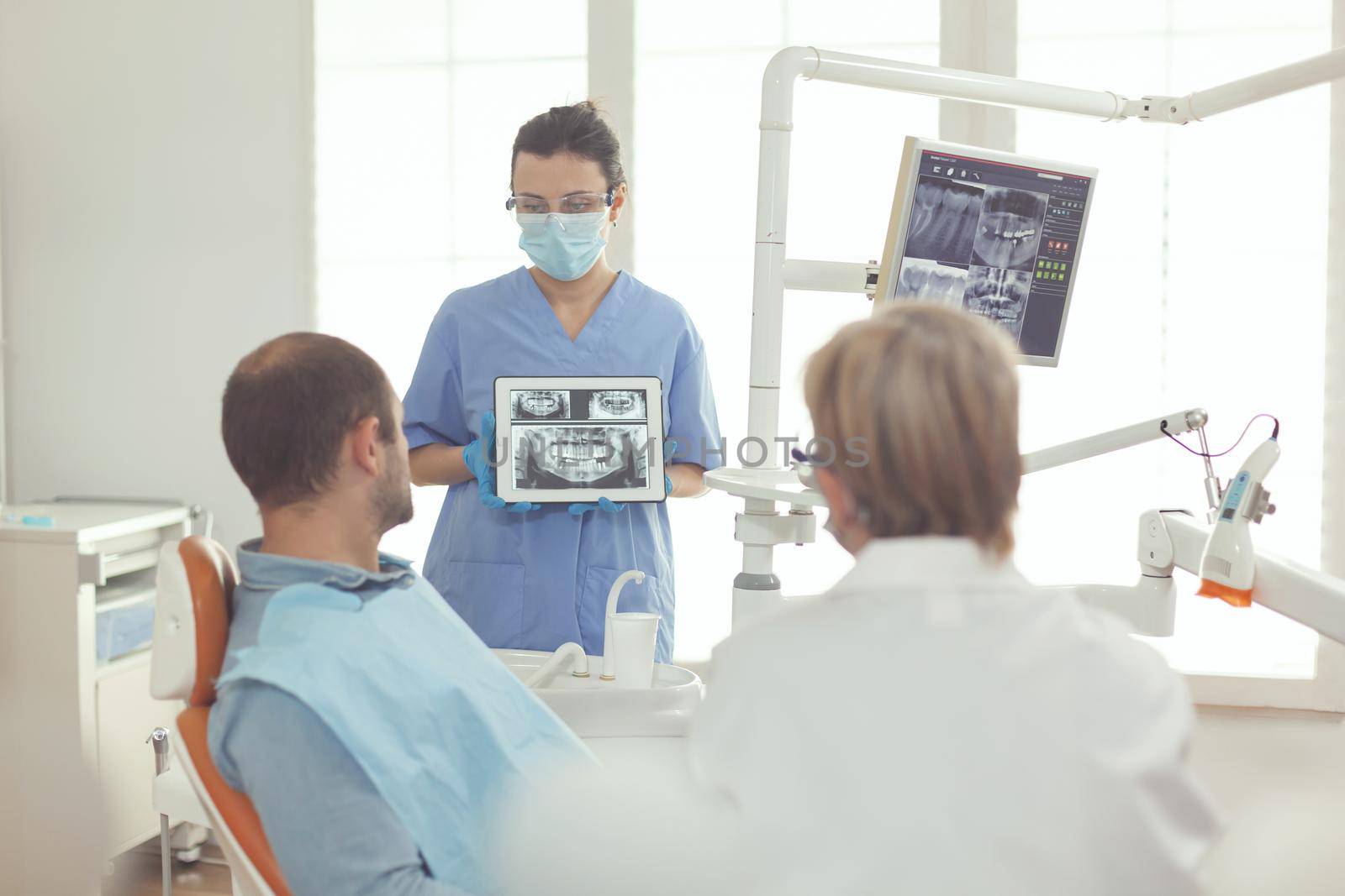 Senior somatology woman talking to sick man about tooth healthcare while sitting in dental chair. Orthodontist nurse explaining toothache pain using digital tablet with denture xray on screen
