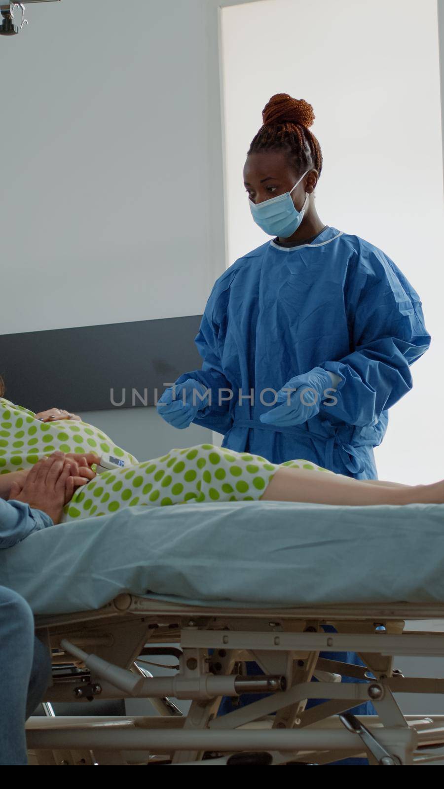 Pregnant person with medical equipment sitting in hospital ward. African american nurse consulting expecting patient in bed and father of child explaining childbirth and delivery procedure