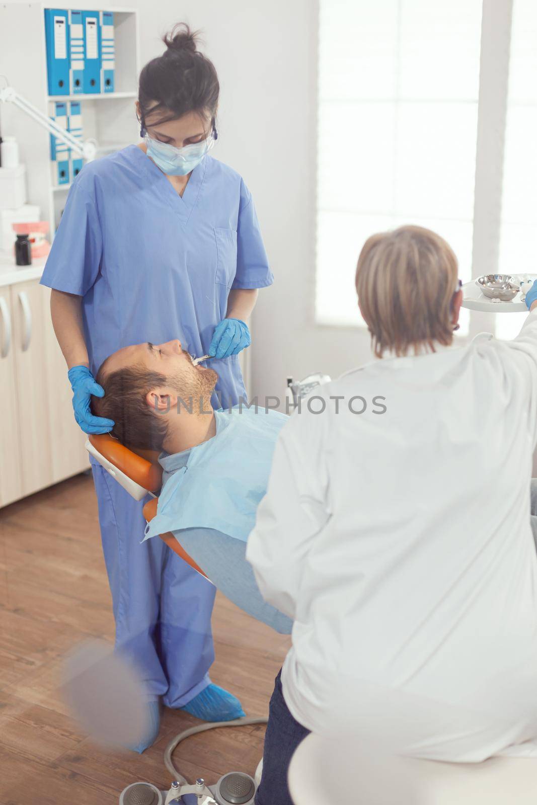 Sick patient standing on stomatology chair with open mouth for dental procedure, senior dentist preparing tools for oral problems. Dentist dentist and medical nurse removes tooth