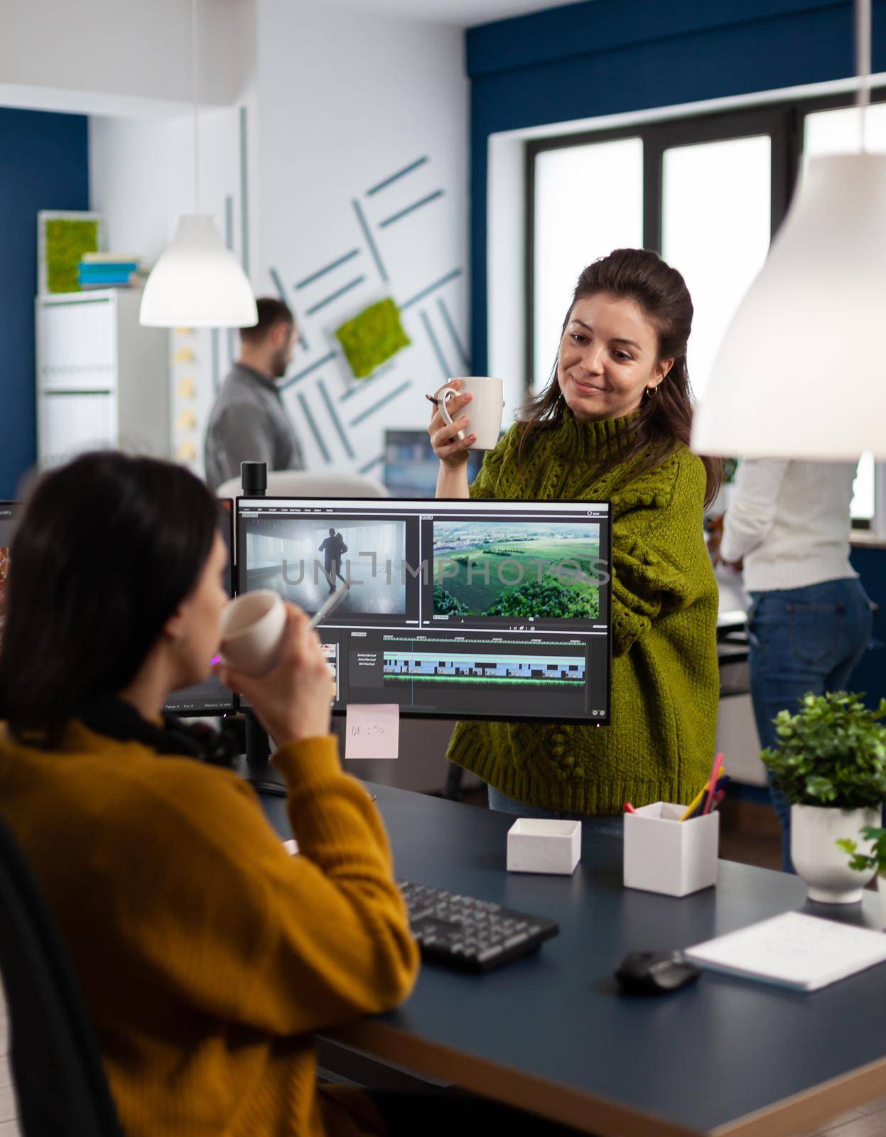 Team of videographers talking about project while drinking coffee working in video editing app at pc sitting in digital multimedia company. Creative women having break while prosessing film montage