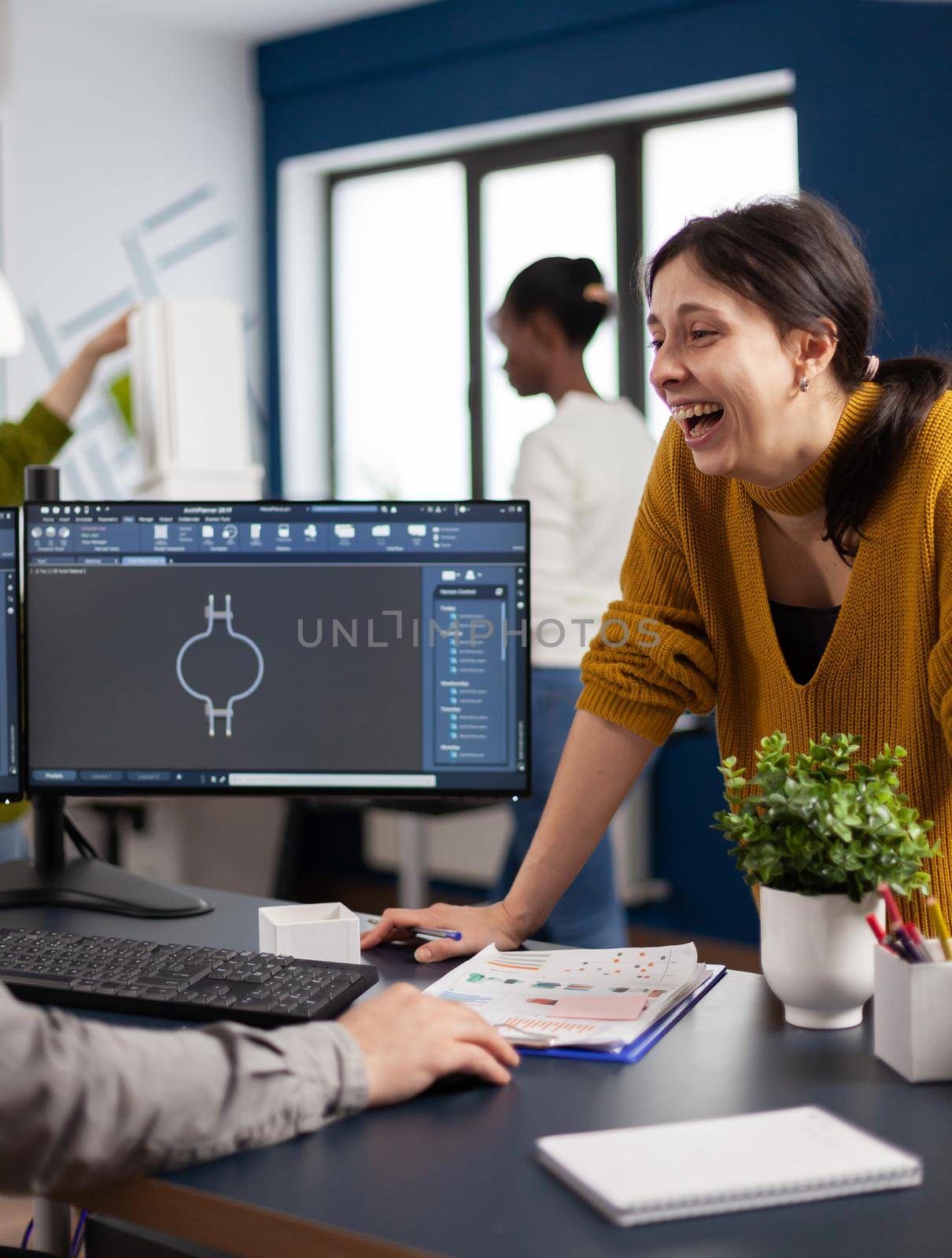 Excited mechanical engineers laughing and discussing about manufacturing new project, taking break. Man technician working at pc, screen shows 3D engine made with CAD software, using computer