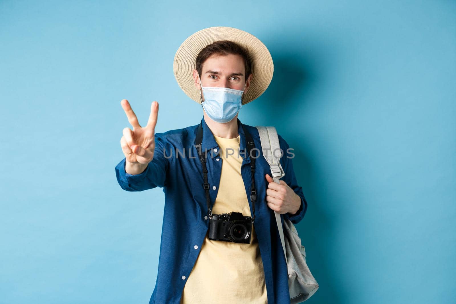 Covid-19, pandemic and travel concept. Positive guy going on vacation, wearing summer hat and medical mask, showing peace sign, standing with backpack and camera on blue background.