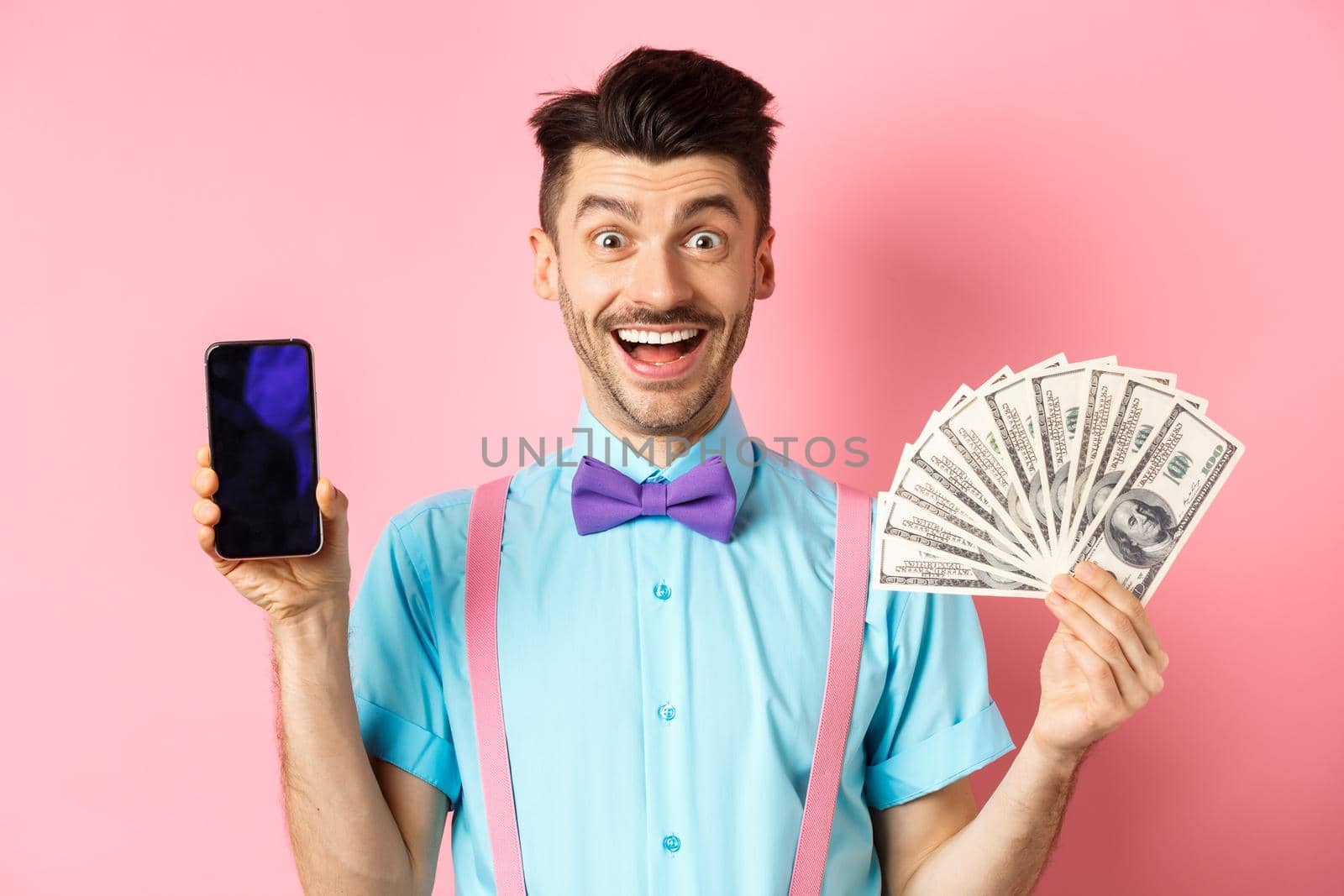 E-commerce and shopping concept. Cheerful guy in bow-tie showing empty smartphone screen and dollars money, smiling amazed at camera, standing on pink background by Benzoix