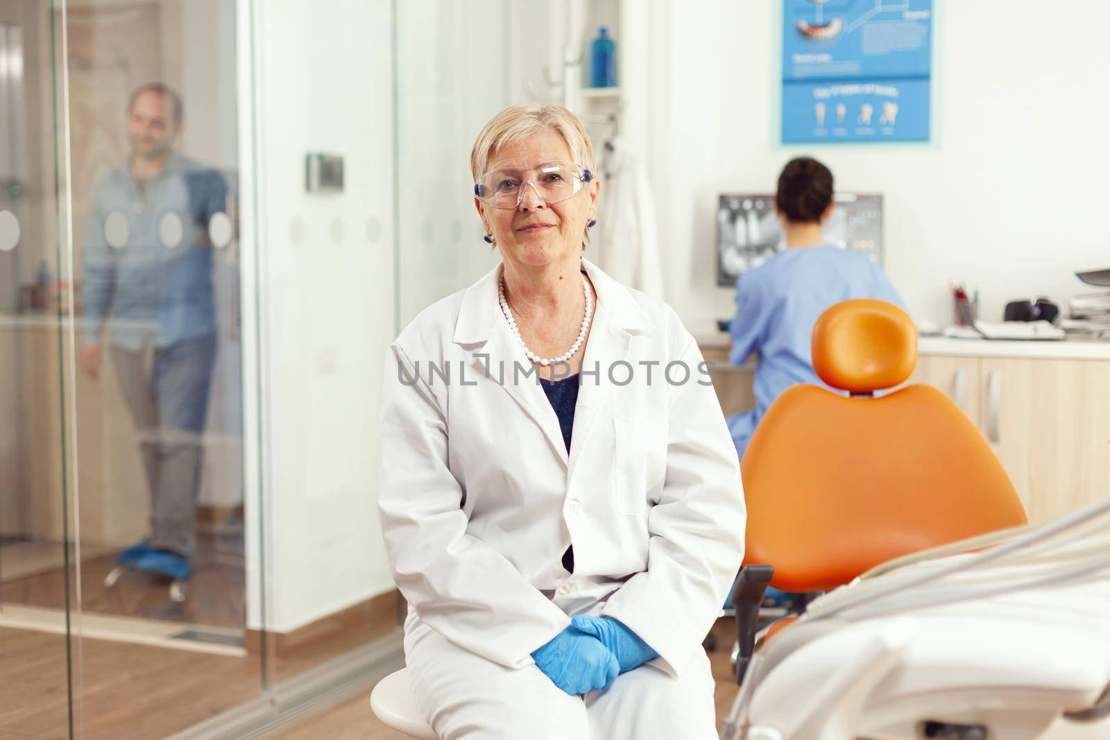 Orthodontist senior in medical uniform sitting on chair looking into camera waiting for man pacient to start stomatology treatment after tooth surgery. Team of dentists working in cabinet office