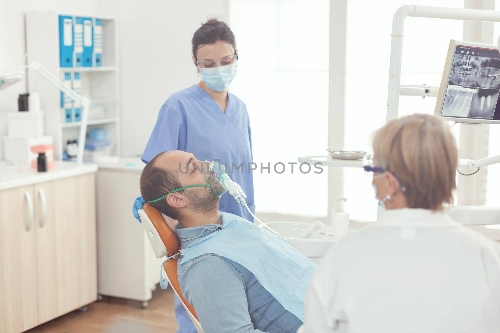 Sick patient sitting on dental chair wearing oxigen mask during stomatology appointment. Dentists hospital team examining tooth while working in dentistry cabinet preparing healthcare treatment