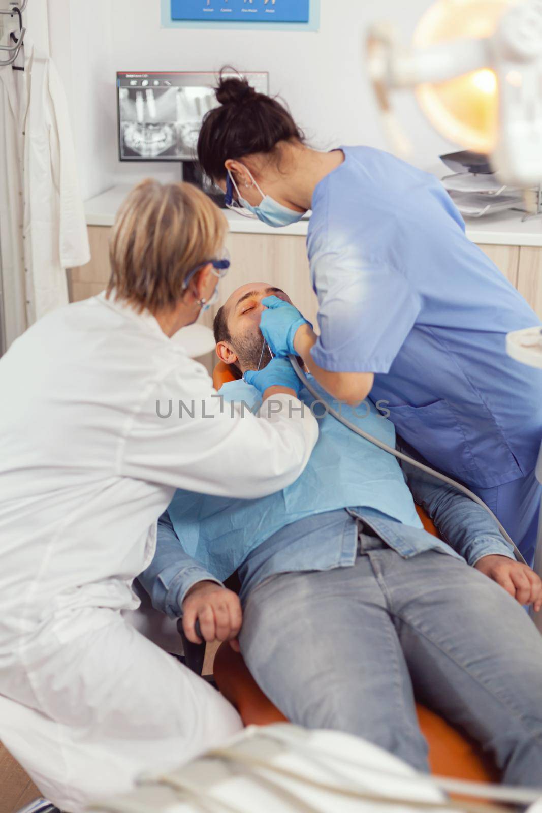 Senior woman technician checking teeth examining sick patient sitting on stomatological chair. Doctor speaking to man while nurse cleaning mouth during dental examination in stomatology clinic