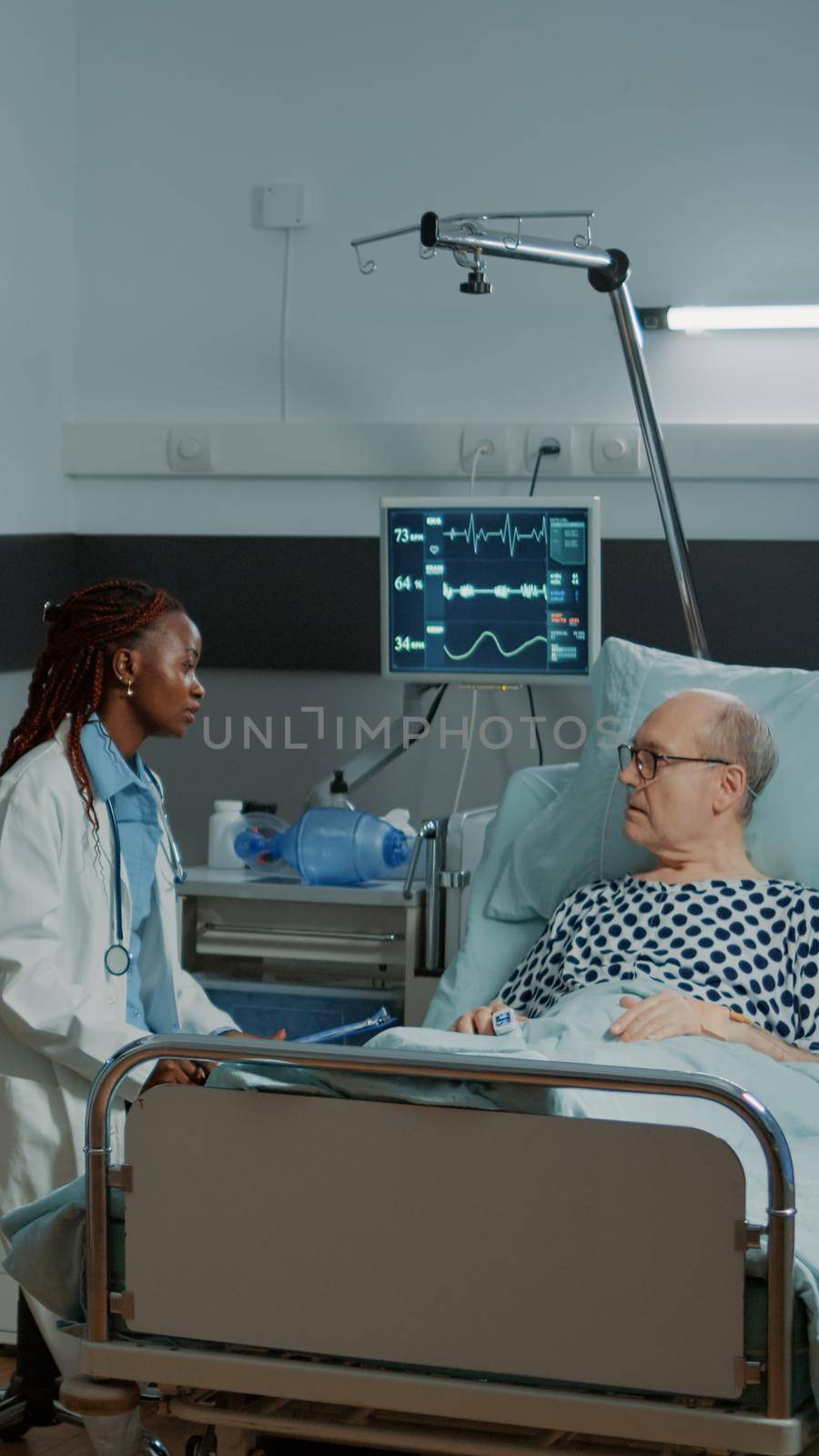 Caucasian nurse checking information files on patient while african american doctor talks to old man feeling sick in modern hospital ward. Multi ethnic medical staff healing disease