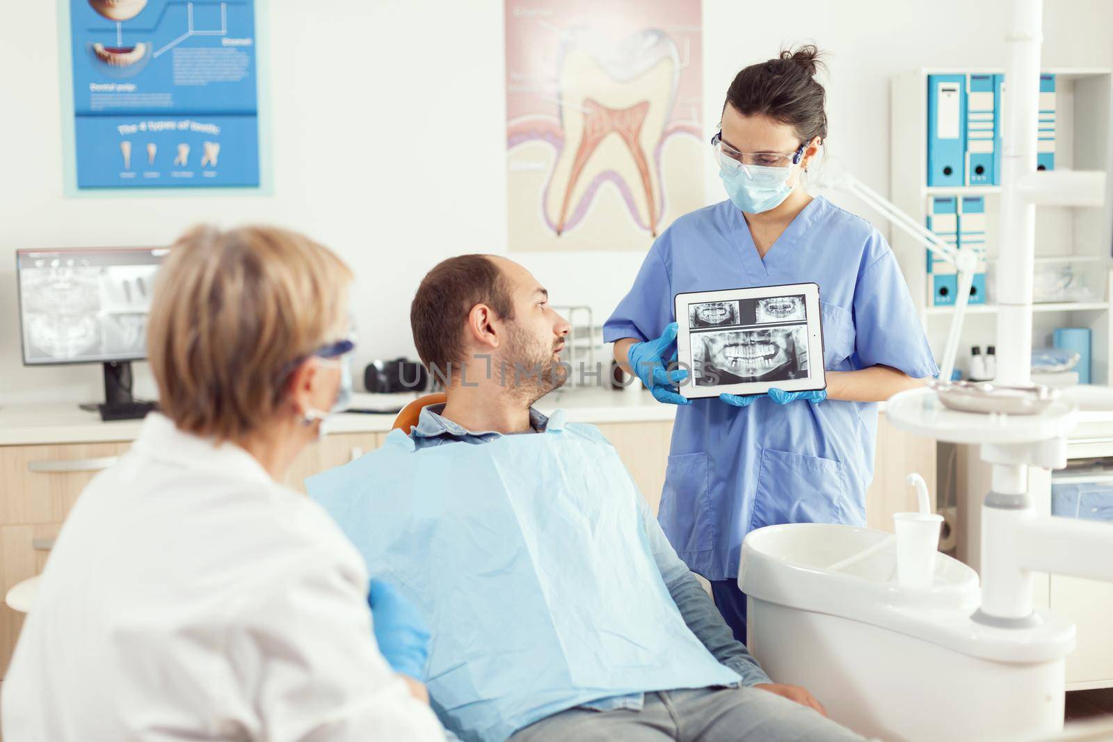 Dentist woman in dental office discussing with sick man while nurse showing tooth x-ray by DCStudio