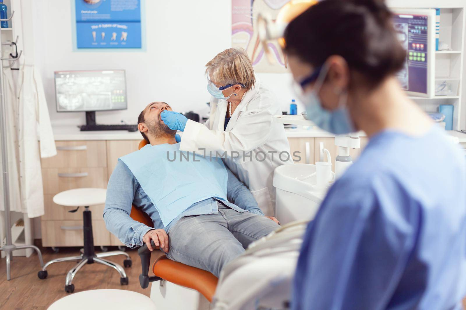 Sick man lying in stomatology chair with open mouth for medical exemination, in orthodontic clinic. Nurse preparing tools for oral problems working in stomatological hospital
