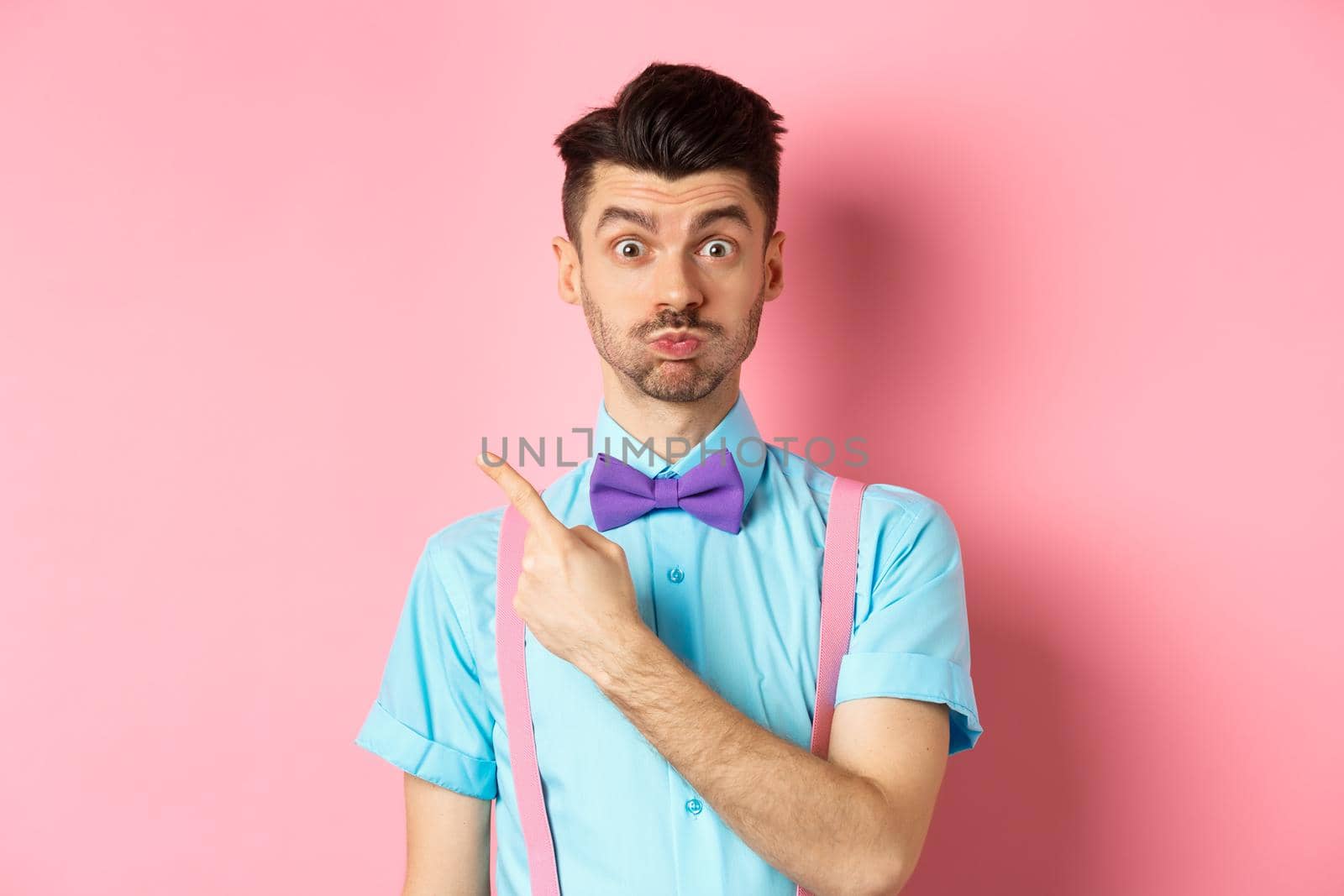 Impressed young man showing awesome promo offer, raising eyebrows and look interested, pointing fingers at upper left corner, pink background.
