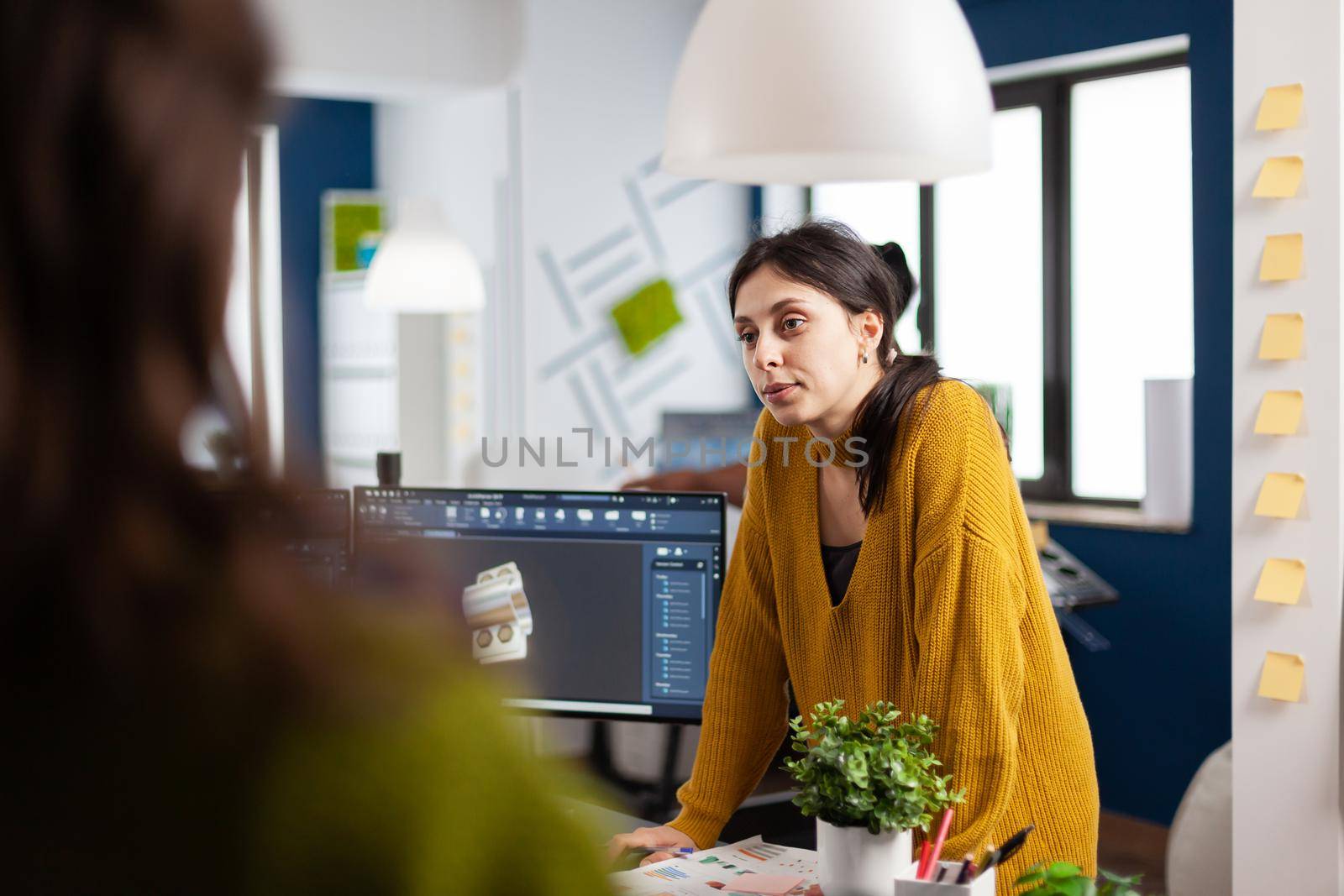 Woman industrial designer discussing with colleague standing at desk by DCStudio
