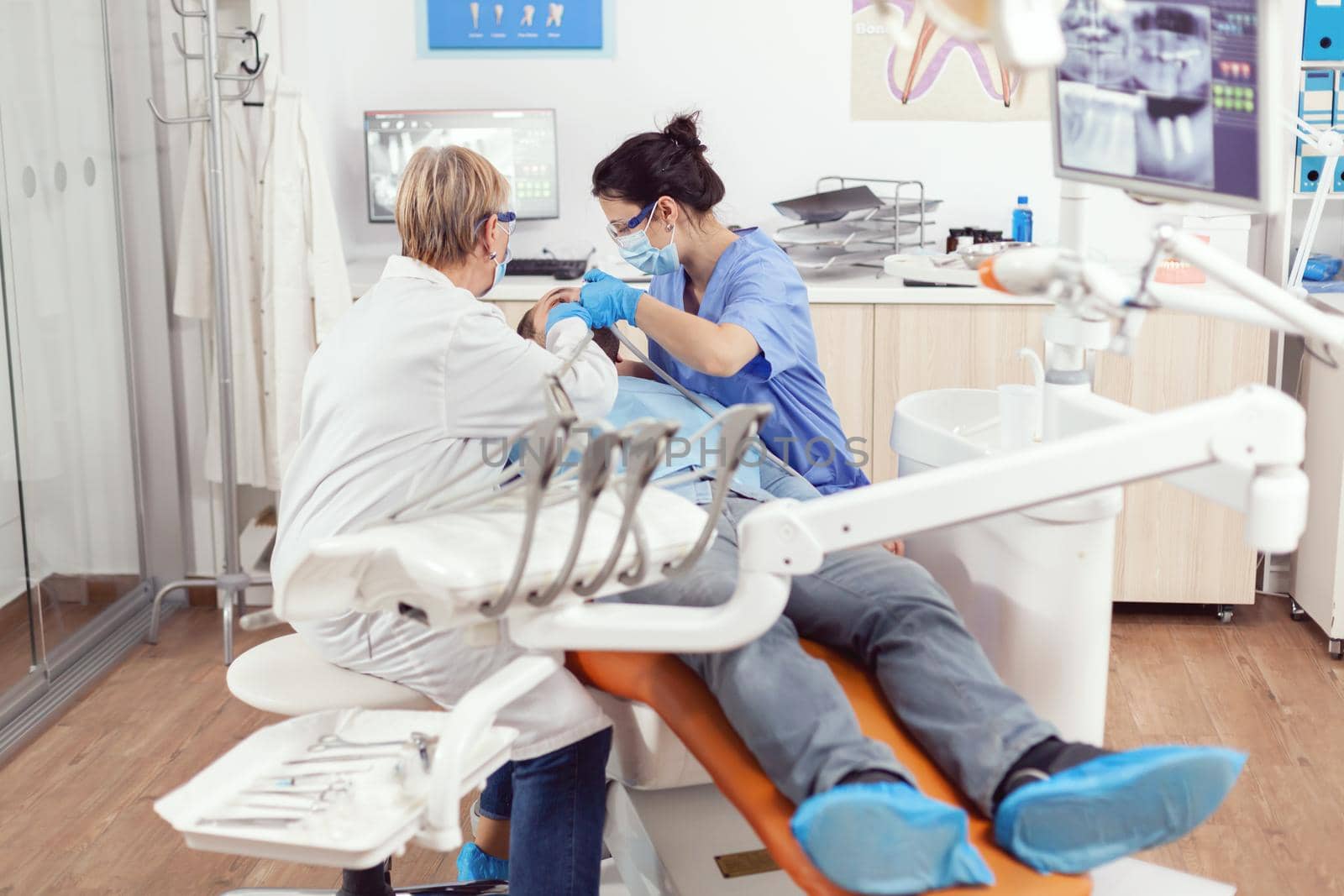 Sick man lying on stomatological chair while senior doctor taking care of tooth health. Orthodontic discussing to patient while nurse cleaning mouth during dental examination in stomatology clinic
