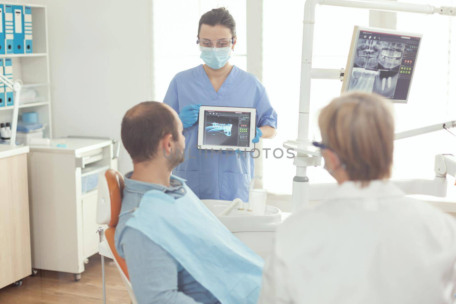 Stomatologist nurse pointing on digital screen explaining tooth x-ray to sick man sitting on dental chair in hospital stomatological office. Dentist doctor discussing toothache treatment