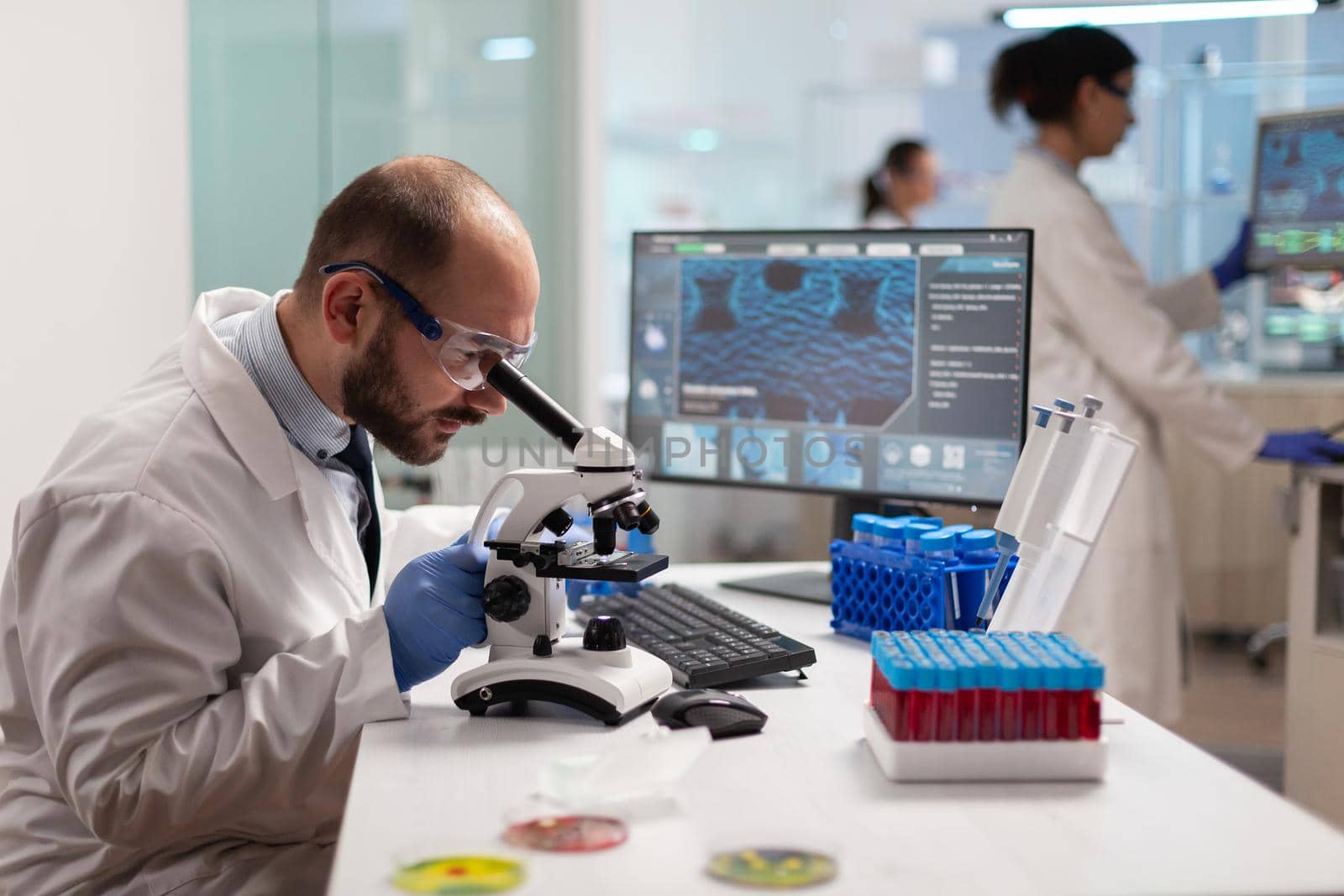 Microbiologist working to find a vaccine for virus in lab looking at sample through microscope weaing protection glasses. Specialists working on medicine, biotechnology research in advanced pharma lab.