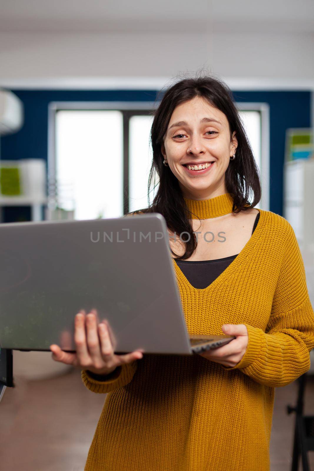 Woman retoucher looking at camera smiling working in creative media agency standing in multimedia company holding laptop. Photographer graphic editor working in start up office at digital assets.
