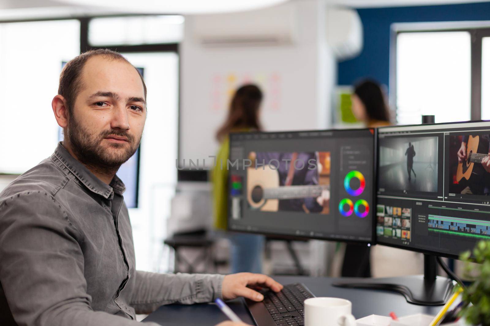 Videographer looking at camera smiling working in creative startup workplace using editing post production software on professional pc with two monitors processing video film montage.