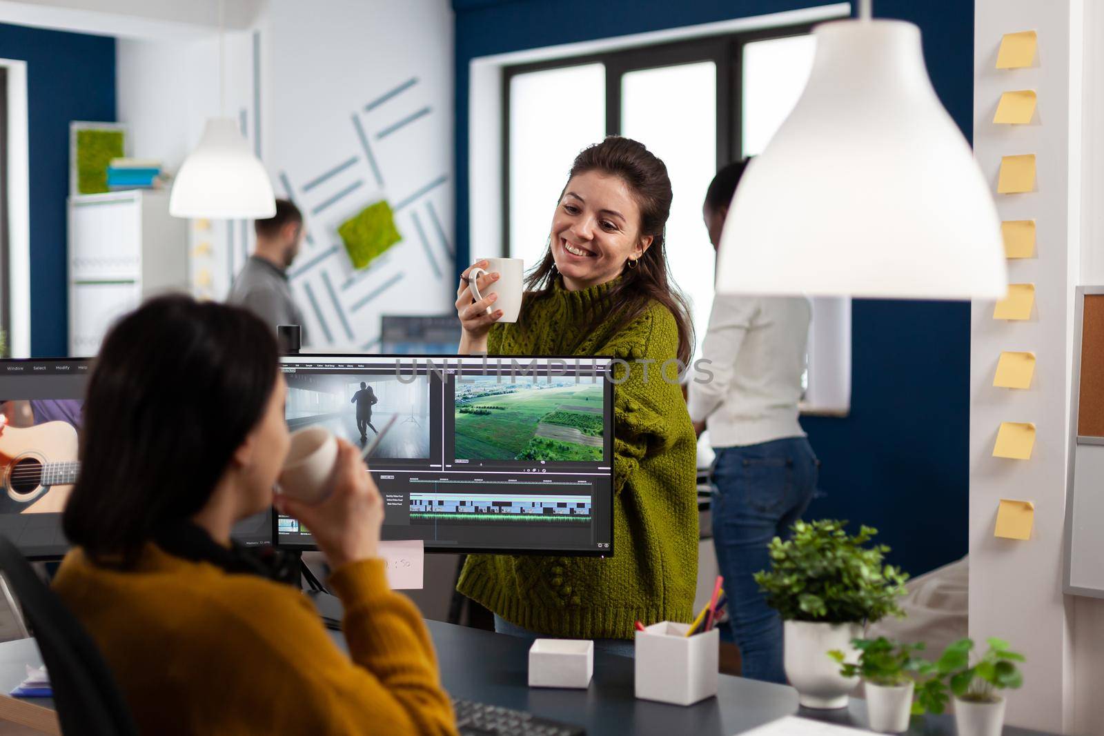 Videographers drinking coffee and talking during work break sitting at desk in creative agency office. Video editor and sound engineer working at project, editing film footage in software app