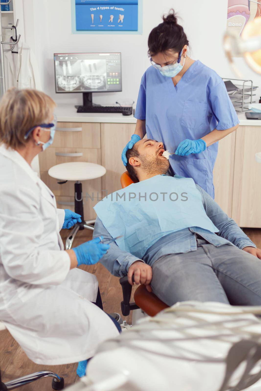 Senior stomatologist woman preparing for surgery putting implant to sick patient mouth in stomatological hospital. Dentist doctor with protection mask treating teeth of man sitting on dental chair