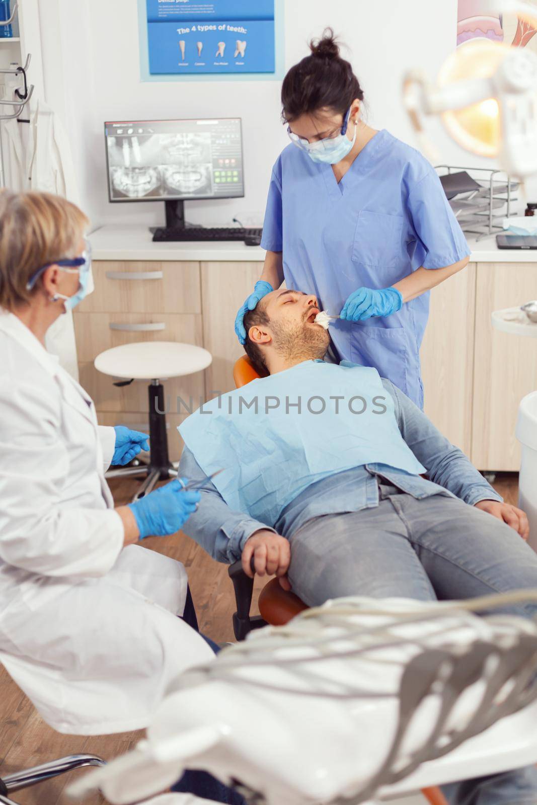 Stomatologist nurse looking in patient mouth preparing for dental surgery. Dentist asisstant with face mask examining sick man with dental bib before intervention sitting on stomatological chair