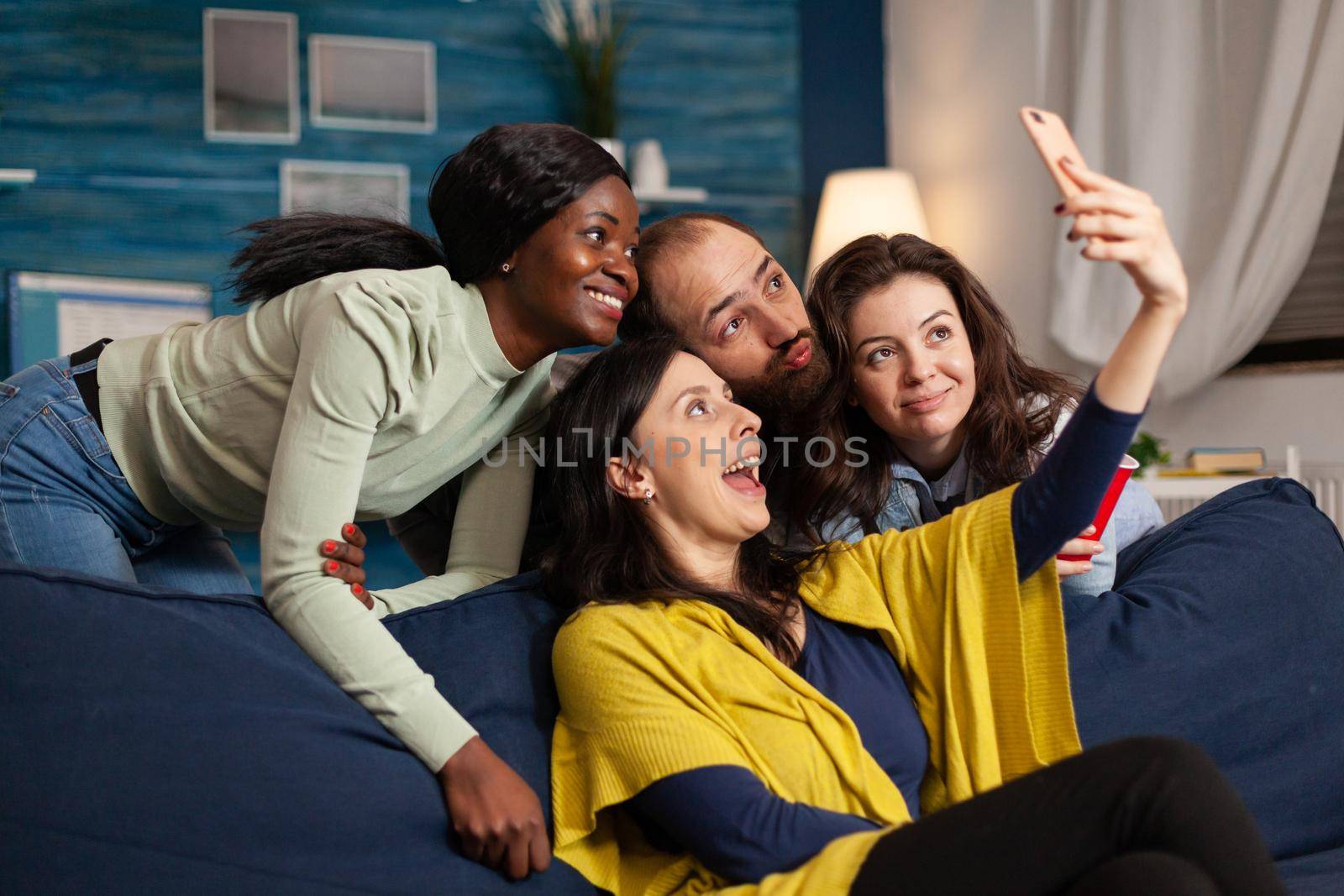 Multiethnic friends making funny faces while taking selfie photo partying in apartment, drinking beer. Group of diverse people laughting, sitting on sofa late at night in living room.