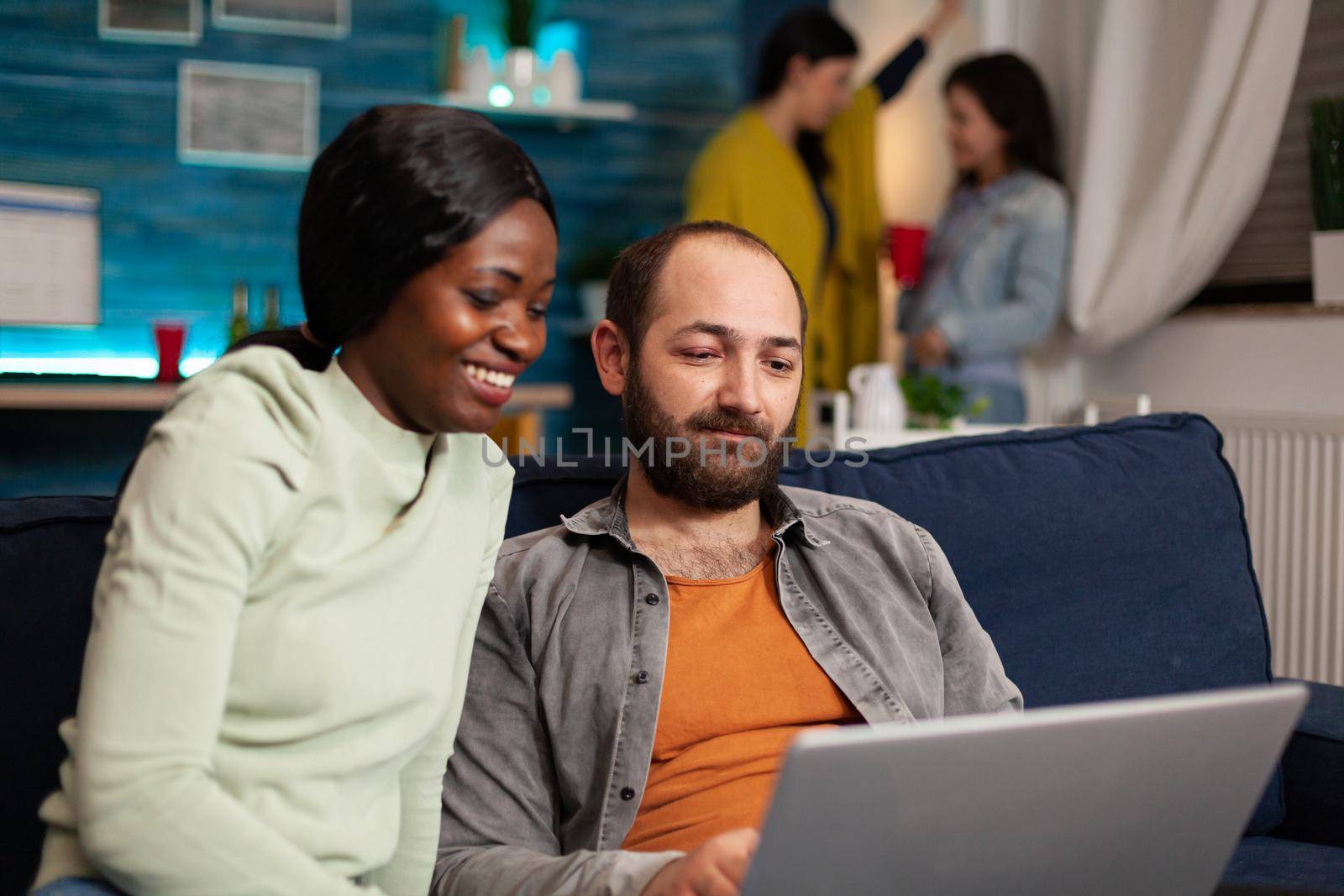 Multiracial friends socializing while watching online videos on laptop relaxing on sofa. In background two women drinking beer enjoying time spend together during entertainment party.