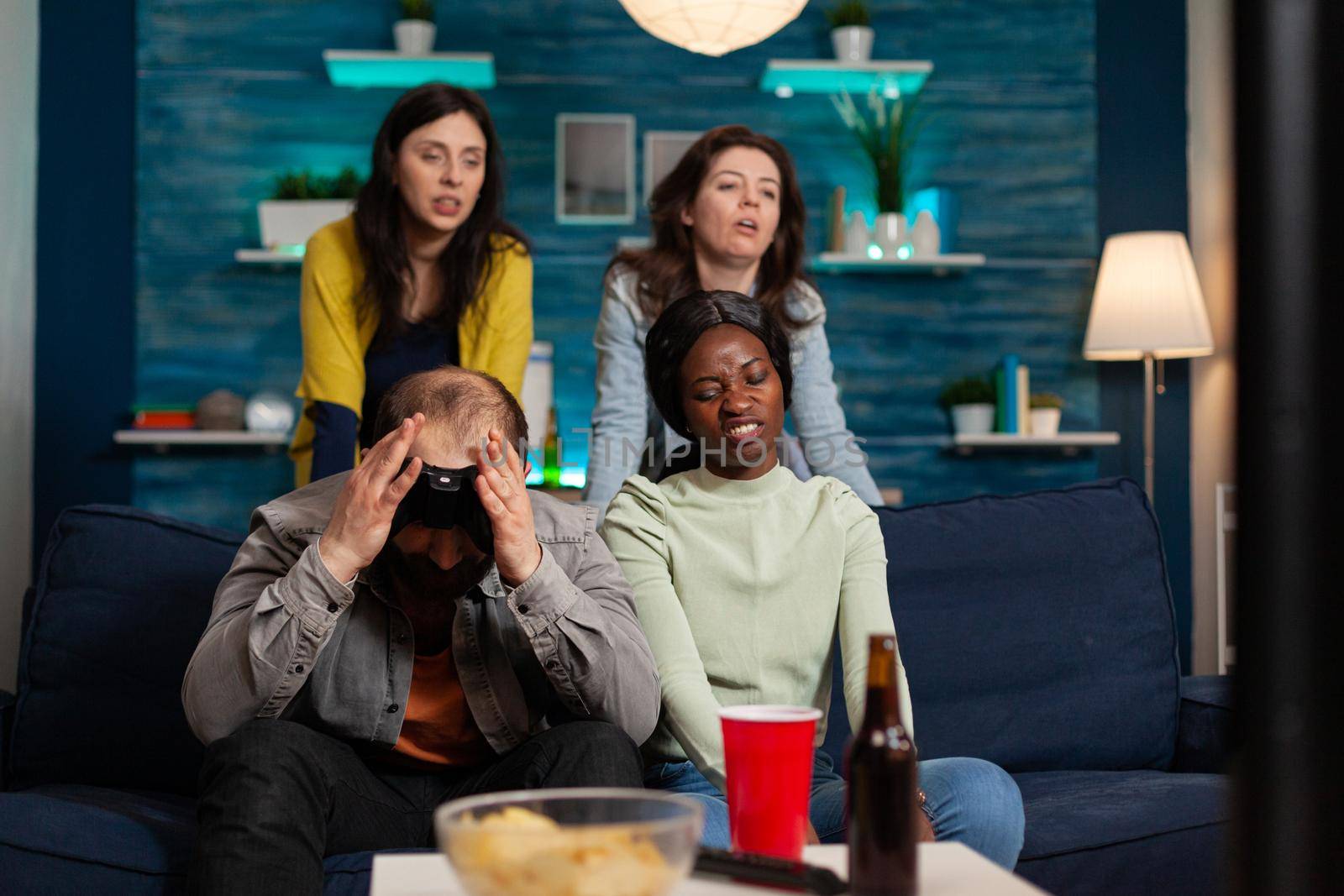 Angry man and multi ethnic friends upset after losing game competition, bonding and sitting on couch after drinking beer. Mixed race group of people hanging out together having fun late at night in living room