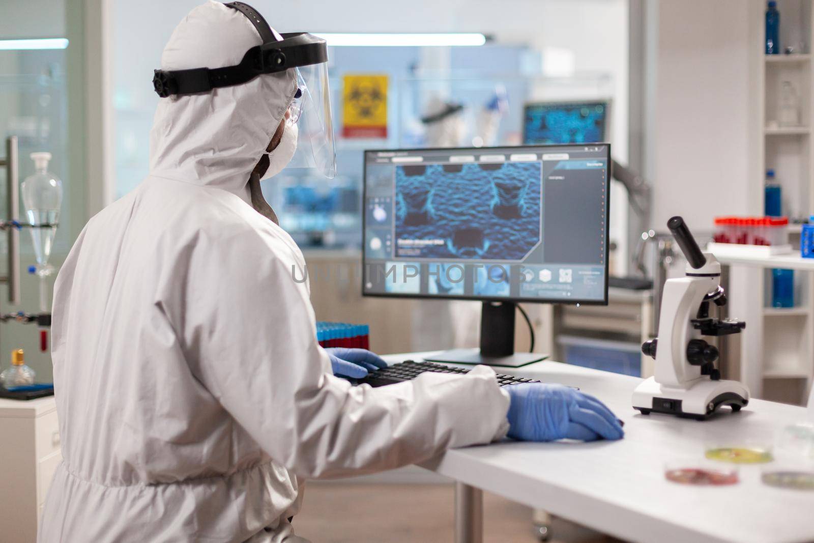 Scientist in protection suit typing on pc working in medical laboratory, Examining vaccine evolution using high tech technology and chemistry tools for scientific research virus development.