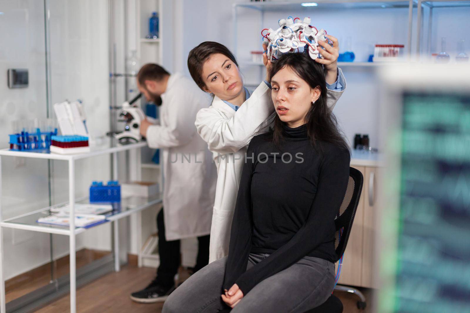 Woman wearing brainwave scanning headset sits in chair in modern laboratory brain study, neurologist looking through microscope in modern medicine lab. Team of doctors using high tech technology.