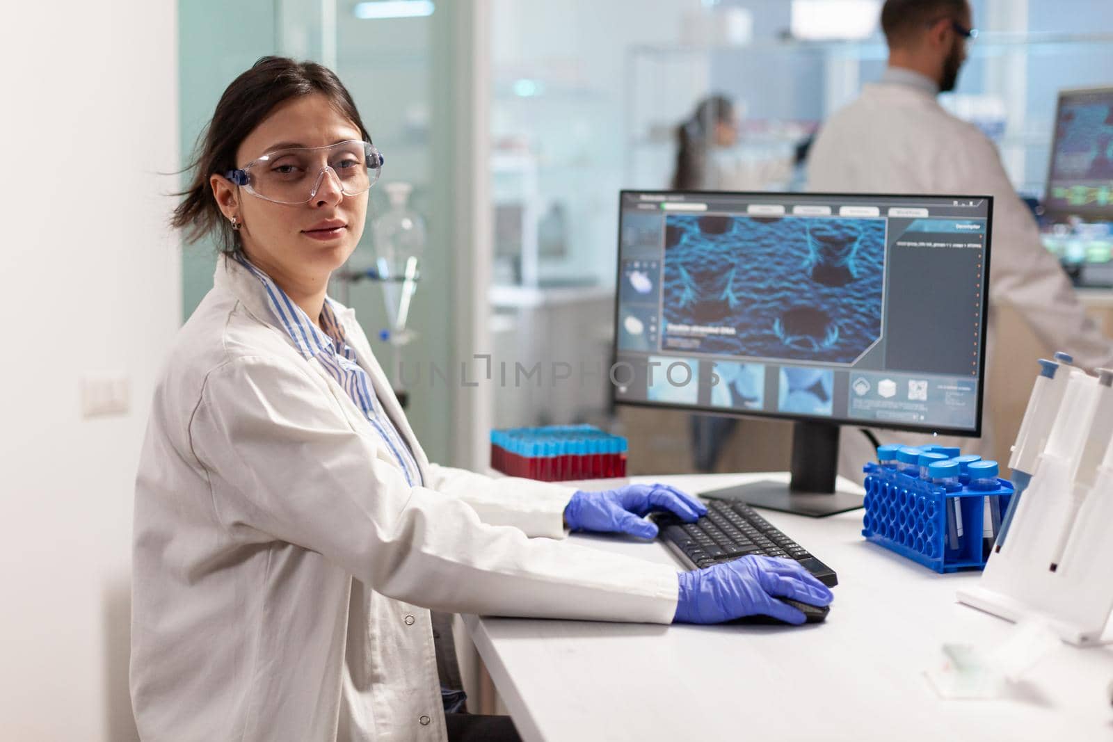 Portrait of scientist looking tired at camera sitting in modern equipped laboratory, typing on pc. Chemist examining virus evolution using high tech for scientific research, vaccine.