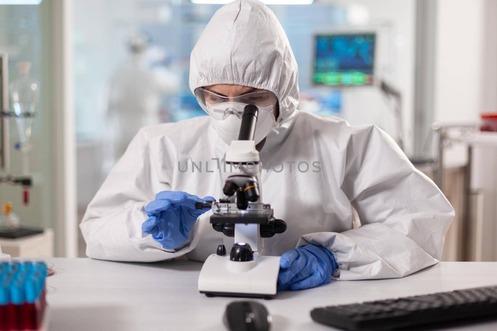 Scientist in ppe suit making adjustments and looking through laboratory microscope. Chemist in coverall working with various bacteria, tissue blood samples for antibiotics research.