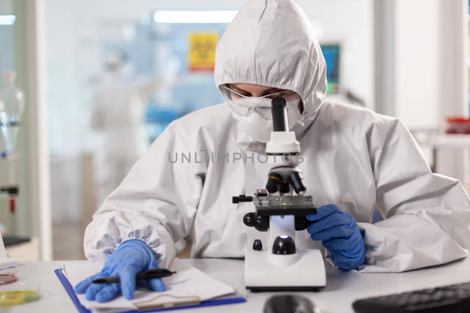 Scientist working to discover virus vaccine dressed in ppe suit looking through microscope. Chemist in coverall working with various bacteria, tissue blood samples for antibiotics research.