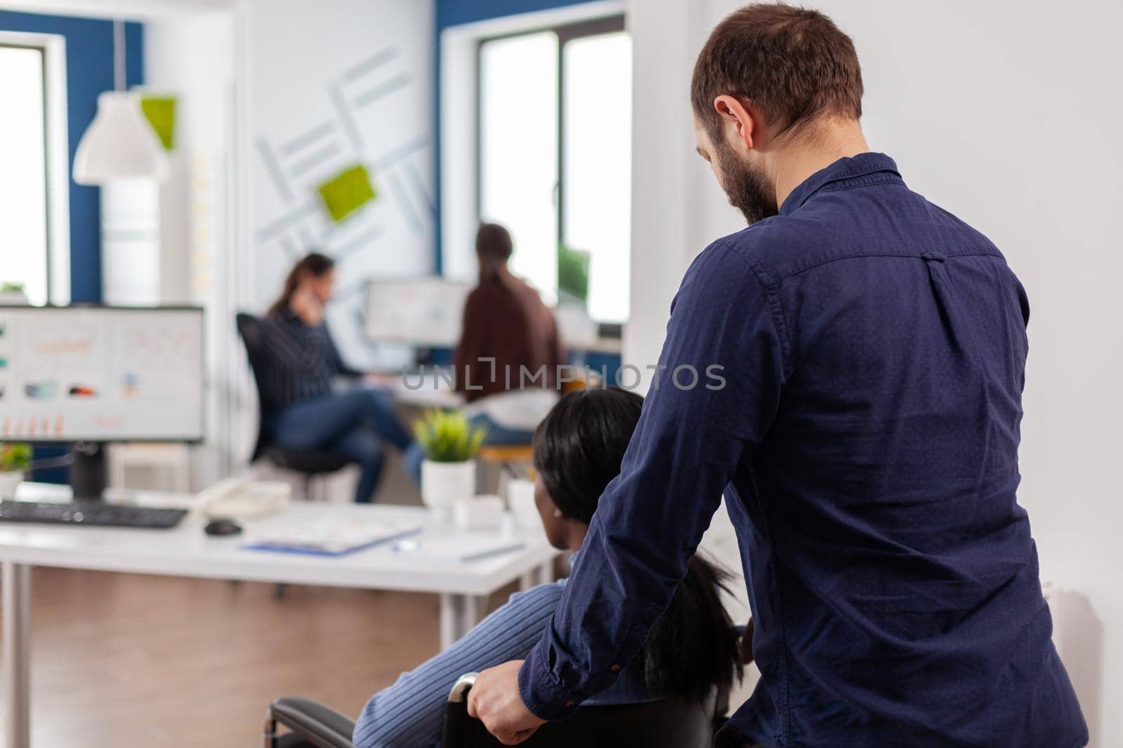 Businessman helping his disabled black colleague entering in company office pushing wheelchair near desk. African handicapped paralysed woman greetting colleagues starting working at financial project.