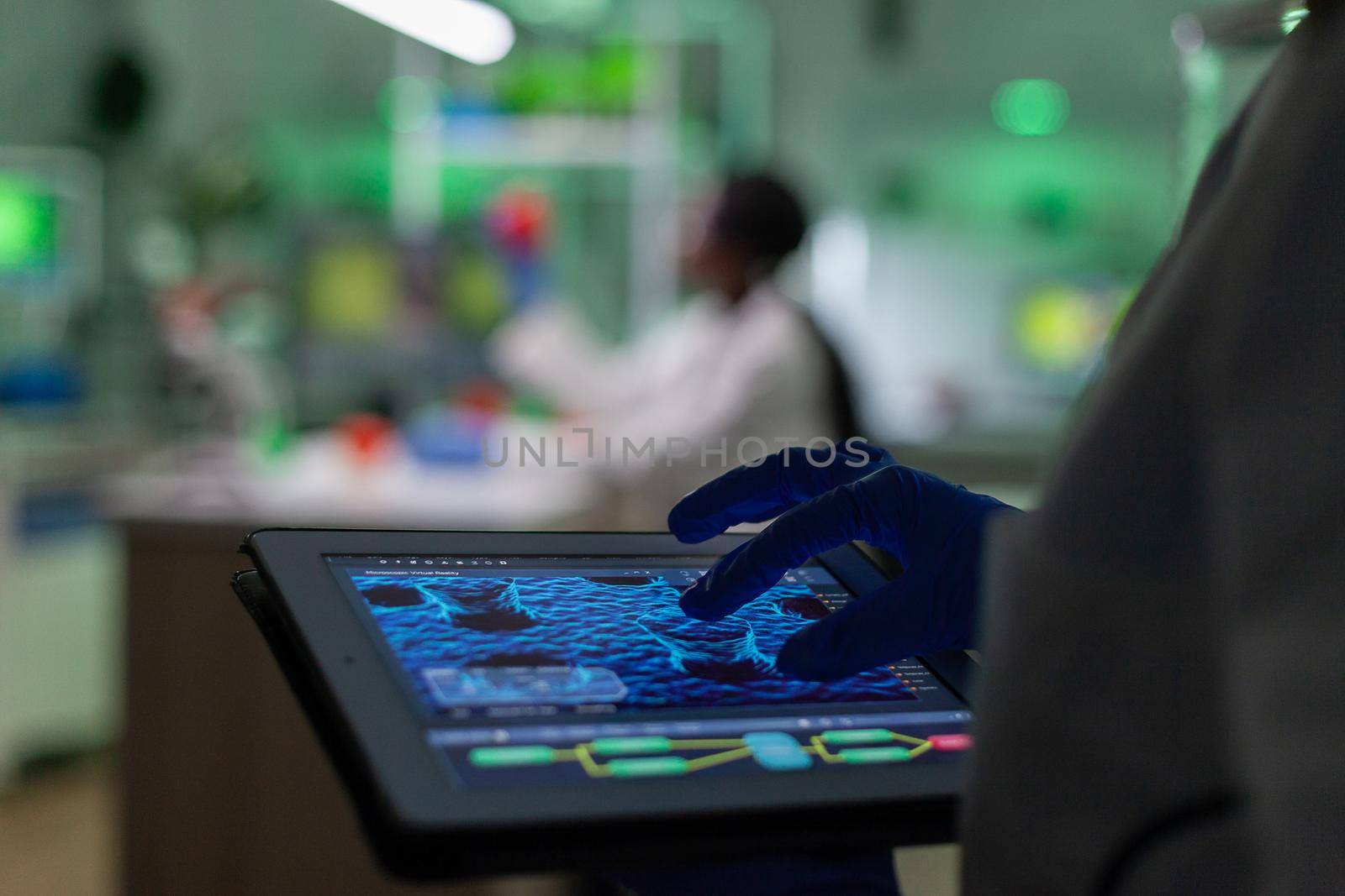 Scientist researcher doctor analyzing genetic mutation test using tablet while her collegue typing biological expertise on computer in background. Medical team working in microbiology laboratory.