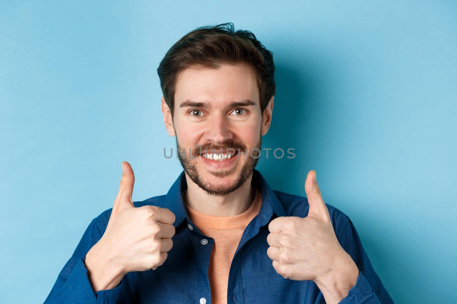 Close-up of smiling good-looking man showing thumbs up, praising good job, recommending company, standing on blue background.