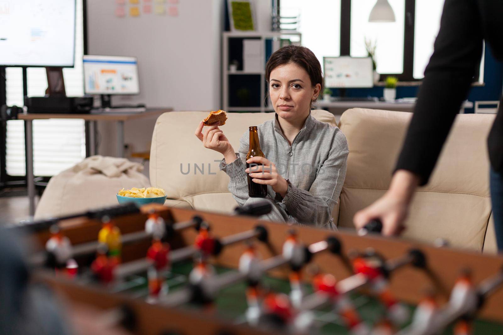 Woman sitting at foosball table at office after work with colleagues eating pizza drinking having fun. Caucasian person watch playing soccer with friends coworkers for entertainment