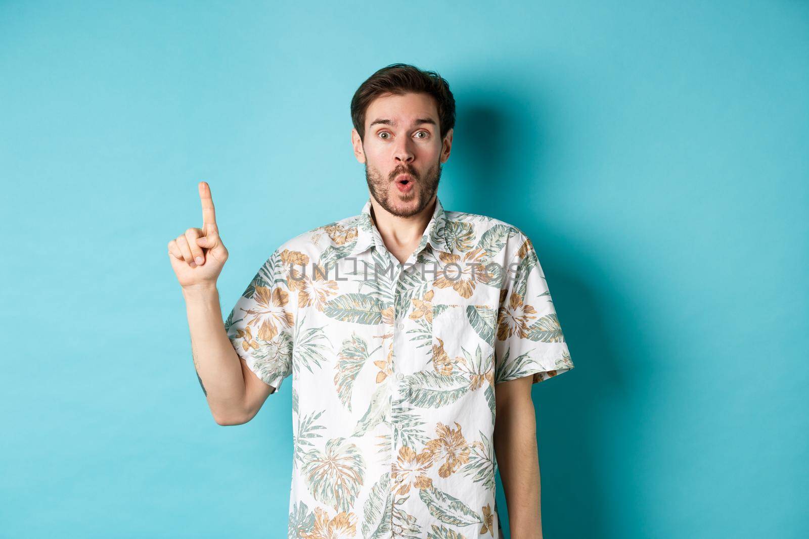 Surprised caucasian tourist in hawaiian shirt, pointing finger up and say wow, checking out promotion, standing on blue background.